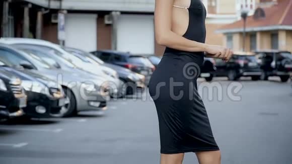 一个穿着时髦黑色连衣裙在停车场摆姿势的苗条女孩从下到上的摄像机布线视频的预览图