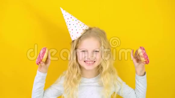 戴着生日帽的疯狂开朗的金发女孩微笑着玩得很开心眼睛上看着两个红色甜甜圈糖果视频的预览图