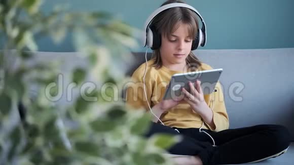 小女孩坐在家里的沙发上一边用耳机和数字平板电脑听音乐视频的预览图