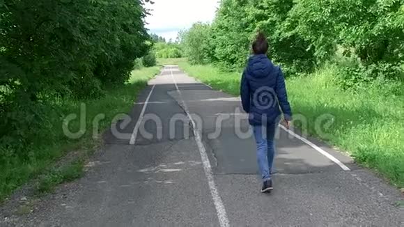 在凉爽的一天穿着蓝色夹克的年轻女子沿着夏日公园的小路散步视频的预览图
