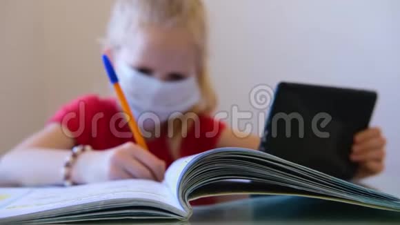 头发模糊的金发女学生戴着医疗面具在家学习做学校作业在笔记本上写作阅读培训视频的预览图