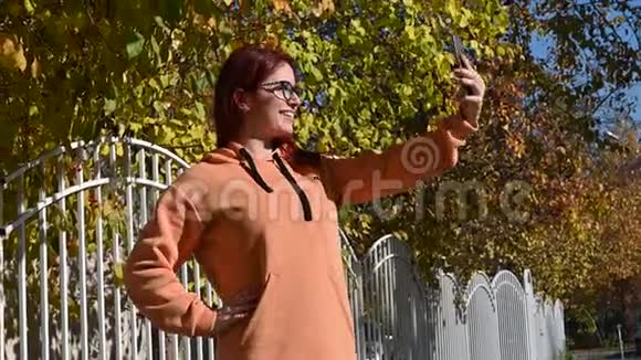 戴眼镜的漂亮红发女人在手机上自拍一个穿着赭色运动衫的快乐女孩被视频的预览图