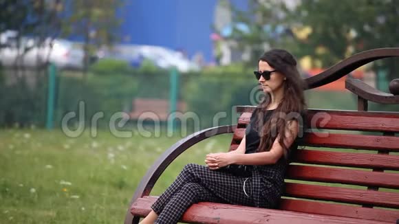 戴墨镜的年轻女子长发坐在公园的长凳上悲伤视频的预览图