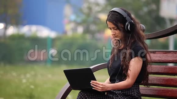 坐在长椅上的年轻女子带着耳机和听音乐的笔记本电脑视频的预览图