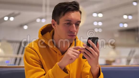 穿着黄色连帽衫的年轻人坐在咖啡馆购物中心使用现代智能手机视频的预览图