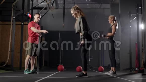 两名女运动员在健身俱乐部用哑铃训练并与教练进行个人锻炼视频的预览图