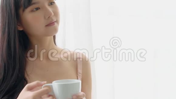 美丽的年轻亚洲女人快乐而自信地喝着一杯咖啡家里有着新鲜而又漂亮的窗户视频的预览图
