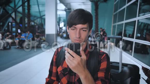 在机场候机楼穿着格子衬衫带行李的微笑男游客使用智能手机等待登机视频的预览图