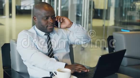 穿着正式服装的非裔美国商人一边看着笔记本电脑一边在智能手机上聊天一边上网视频的预览图