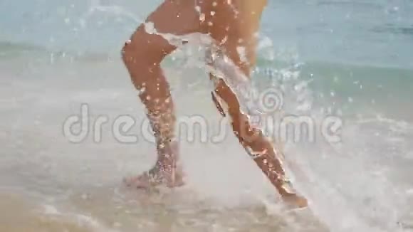 轻柔的小波浪溅在年轻女子沿着海滨奔跑的腿上女性赤脚在浅沙中慢跑视频的预览图