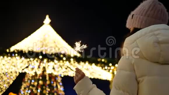穿着外套戴着帽子的年轻女子在圣诞树旁闪闪发光从后面看视频的预览图