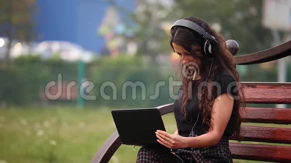年轻女子坐在长凳上带着耳机和笔记本电脑在户外听音乐视频的预览图
