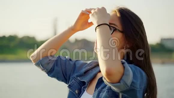 一个戴着眼镜和牛仔夹克的女孩走在海湾边把她的头发弄直了风吹得乌黑的头发闪闪发光视频的预览图