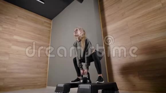 运动女孩正在健身大厅表演蹲手里拿着水壶铃站在平台上视频的预览图