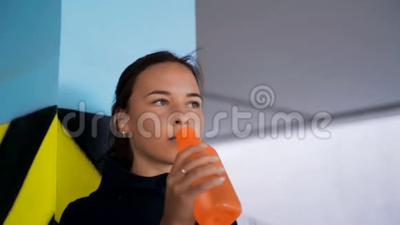 一个穿着黑色夹克的女孩慢跑后喝着橙色瓶子里的水而站在靠近柱子的停车场里视频的预览图