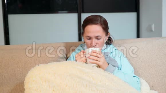 生病的女人感冒了冬天喝热茶柠檬坐在家里的沙发上一个戴着医用口罩的女人视频的预览图