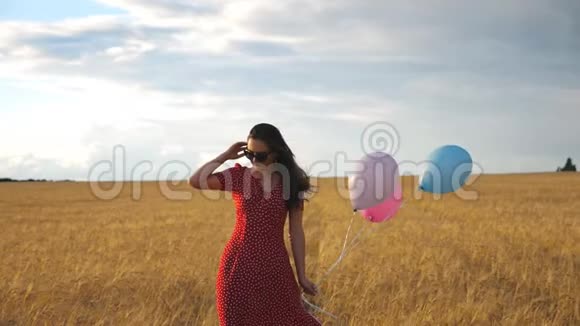 快乐美丽的女孩戴着太阳镜穿过麦田手里拿着气球穿红色衣服的年轻女子视频的预览图
