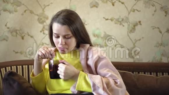 一个漂亮体贴的女孩坐在沙发上喝茶她肩膀上有棕色的毯子妇女视频的预览图
