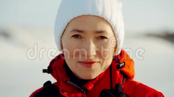 一个穿着红色夹克和白色帽子的女人看着相机冬日阳光明媚概念寒假家庭视频的预览图