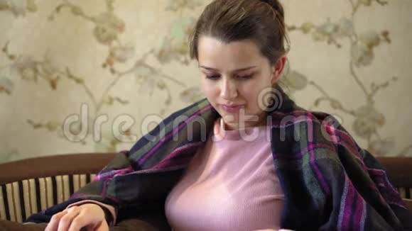 漂亮体贴的女孩坐在沙发上看书的平均计划她的肩膀上有一条棕色的毯子视频的预览图