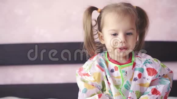 一个穿着晨衣的小女孩坐在沙发上咬着嘴唇的特写镜头有两个辫子的可爱宝宝视频的预览图