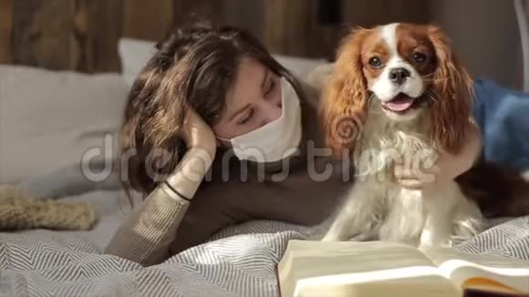 在隔离期间一个年轻的保护性蒙面女孩和她可爱的小狗在一本书旁边的床上缓慢移动冠状病毒视频的预览图
