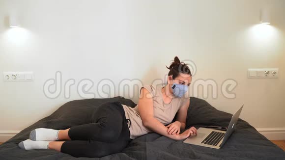 年轻的女人在家里的床上戴着面罩在笔记本电脑上工作视频的预览图