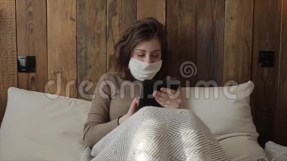 在隔离期间一个戴着防护面罩手中拿着手机的年轻欧洲女孩坐在家里的床上冠状病毒视频的预览图