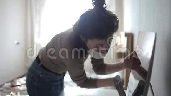 一个戴口罩的年轻欧洲女孩在检疫期间用防腐剂仔细擦拭她的电脑冠状病毒COVID19视频的预览图