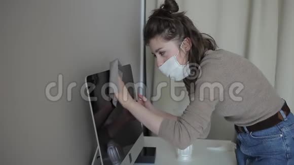 一个在家被隔离的年轻女孩戴着医用口罩用消毒剂仔细擦拭电脑冠状病毒COVID视频的预览图