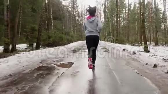 穿着灰色帽衫和黑色运动裤的布鲁内特在森林线的沥青路上跑步后景视频的预览图