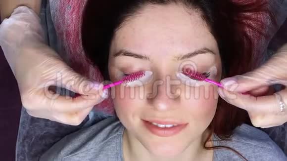 一个女人躺着闭着眼睛做美容手术主人用粉红色的刷子梳理延伸的睫毛顶级视频的预览图