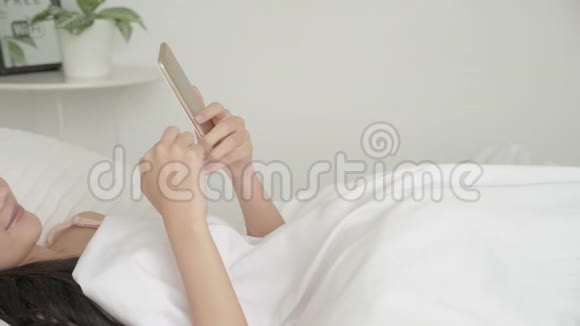 美丽的亚洲年轻女性看着摸着智能手机在卧室的床上享受社交网络视频的预览图
