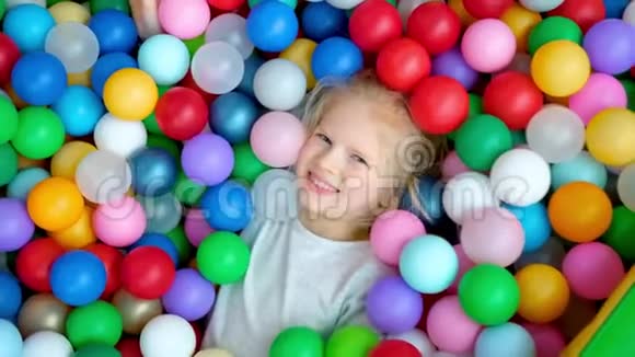 可爱的金发小女孩躺在多种颜色的塑料球在大干划水池在游戏中心微笑着视频的预览图