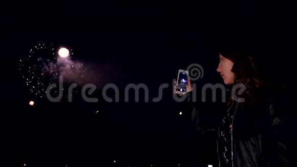 夜晚的女孩看着天空中的烟花在智能手机上制作一段视频视频的预览图