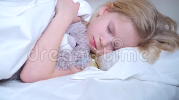 睡着的小女孩在睡梦中抱着一只泰迪熊一个金发碧眼的孩子早早地睡在床上铺着白色床上用品视频的预览图