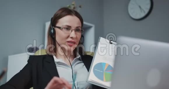 耳机里的女商人在网络摄像头上写财务报告在办公室打视频电话商务教练视频的预览图