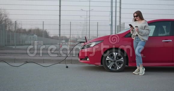 时髦的女孩站在她的红色电动车旁边等待车辆充电连接充电器视频的预览图