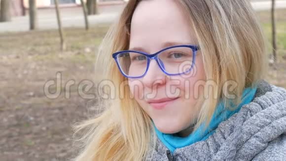 一位金发碧眼戴着眼镜坐在春天公园里微笑的年轻美女的肖像视频的预览图