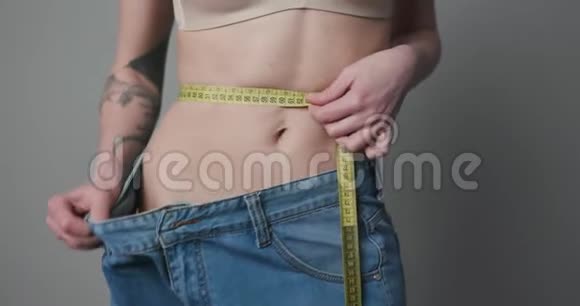 年轻女人穿着大牛仔裤减肥概念女人用胶带控制她的措施女人在测量身体视频的预览图