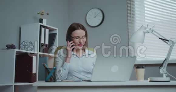 年轻的休闲女士坐在办公桌前用笔记本电脑接电话打电话微笑着在手机上说话视频的预览图