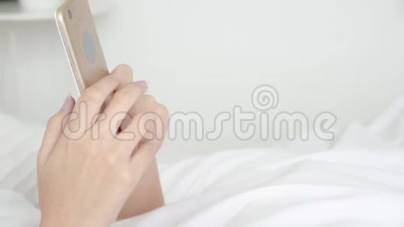 美丽的亚洲年轻女性看着摸着智能手机在床上享受社交网络视频的预览图