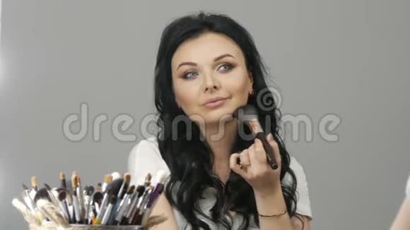 一个漂亮的年轻女人在镜子前有一个长长的黑色卷发用一种特殊的化妆刷涂着腮红视频的预览图