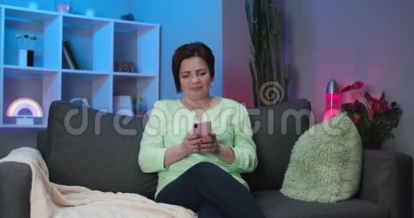 一位留着黑色短发坐在家里的沙发上手里拿着看不见的智能手机和聊天的白人女性视频的预览图