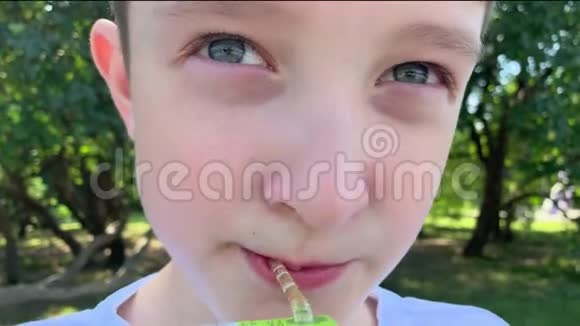一个十几岁的白种人小男孩坐在夏天的城市公园里用纸汁袋里的吸管喝果汁视频的预览图
