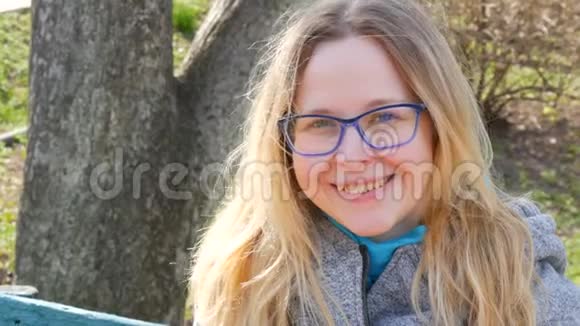 美丽的年轻女子金发碧眼戴着眼镜坐在春天的公园里微笑视频的预览图