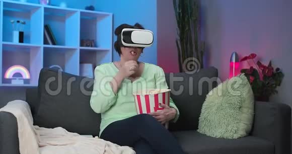 老年妇女戴着虚拟现实眼镜一边吃爆米花一边坐在沙发上在家看电影视频的预览图