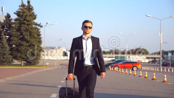 英俊的年轻商人戴着太阳镜走在城市街道上把手提箱拉在轮子上自信的成功人士视频的预览图