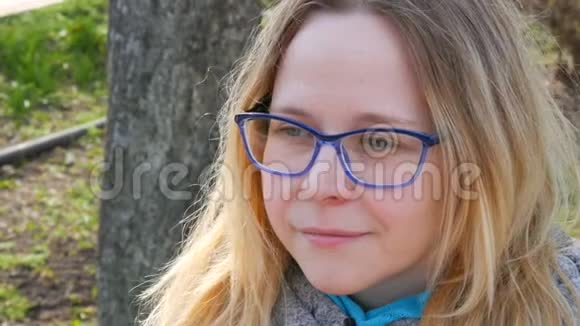 一位金发碧眼戴着眼镜的美丽年轻女子的肖像坐在一个春天的公园里微笑视频的预览图