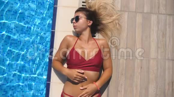 美丽的女孩戴着太阳镜和红色比基尼躺在酒店游泳池附近用手触摸水面视频的预览图